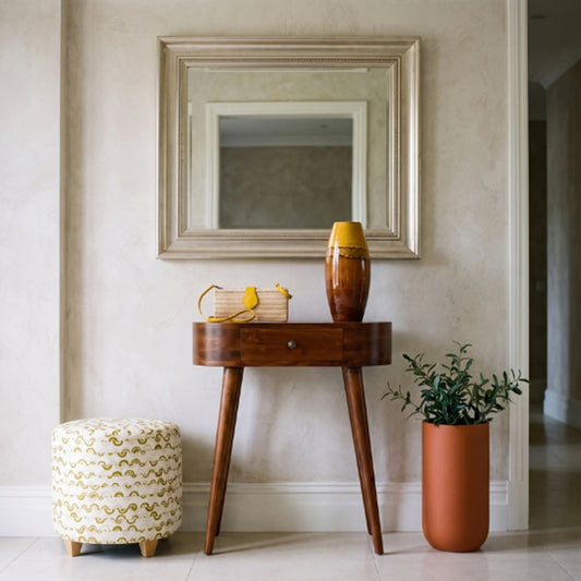 Chestnut oval solid wood console table with a single drawer and tapered legs, styled beneath a wall mirror beside a patterned stool and potted plant.