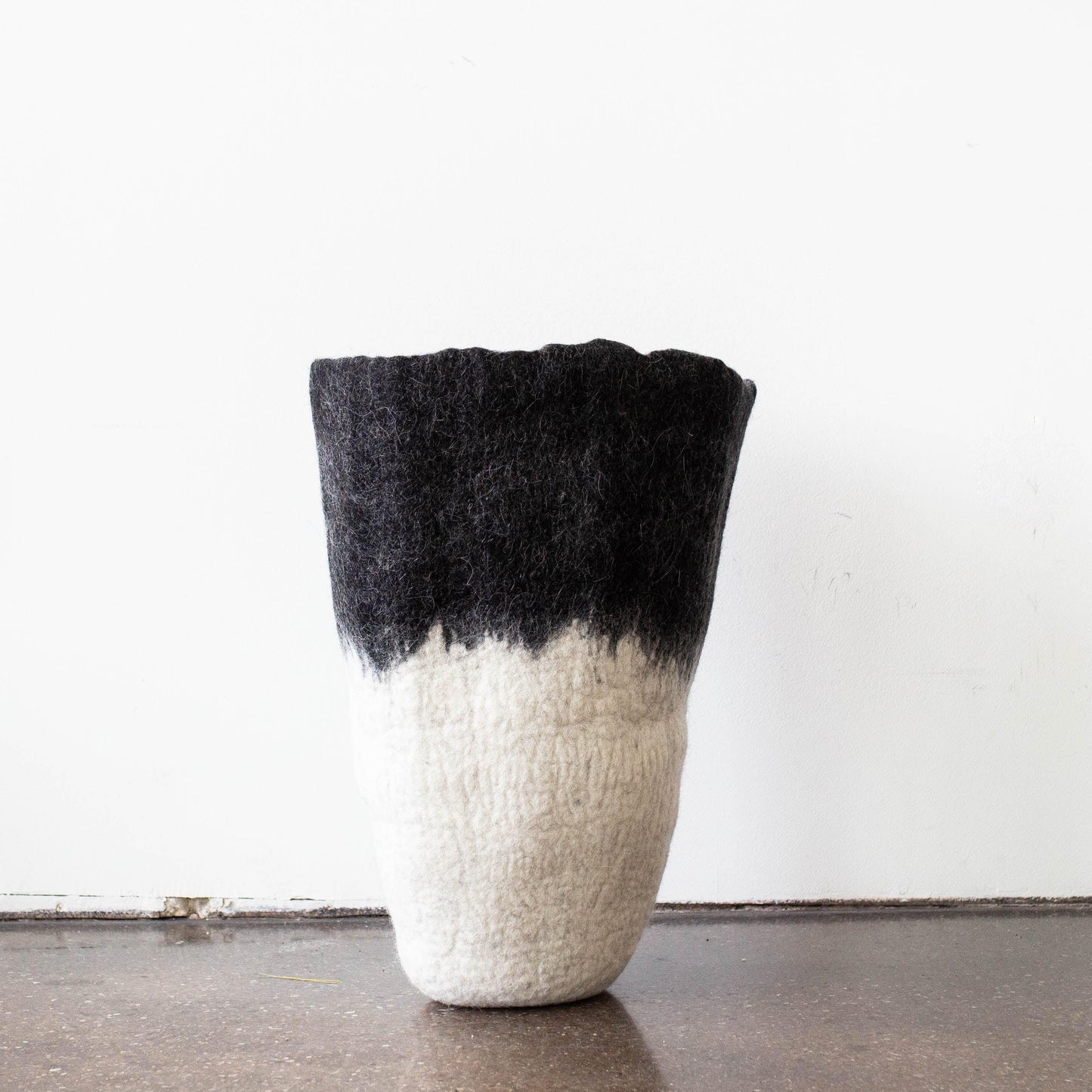 Sculptural black to white ombré felted wool floor vase standing alone on dark wood floor against clean white wall

