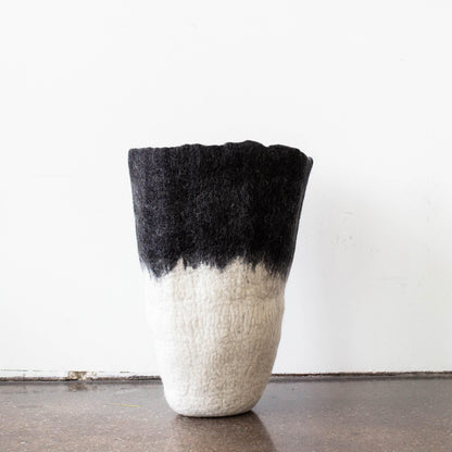 Sculptural black to white ombré felted wool floor vase standing alone on dark wood floor against clean white wall

