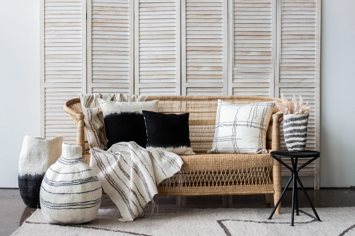 Black and white ombré wool floor vases styled beside rattan sofa with neutral throw pillows and woven blanket against whitewashed shutters

