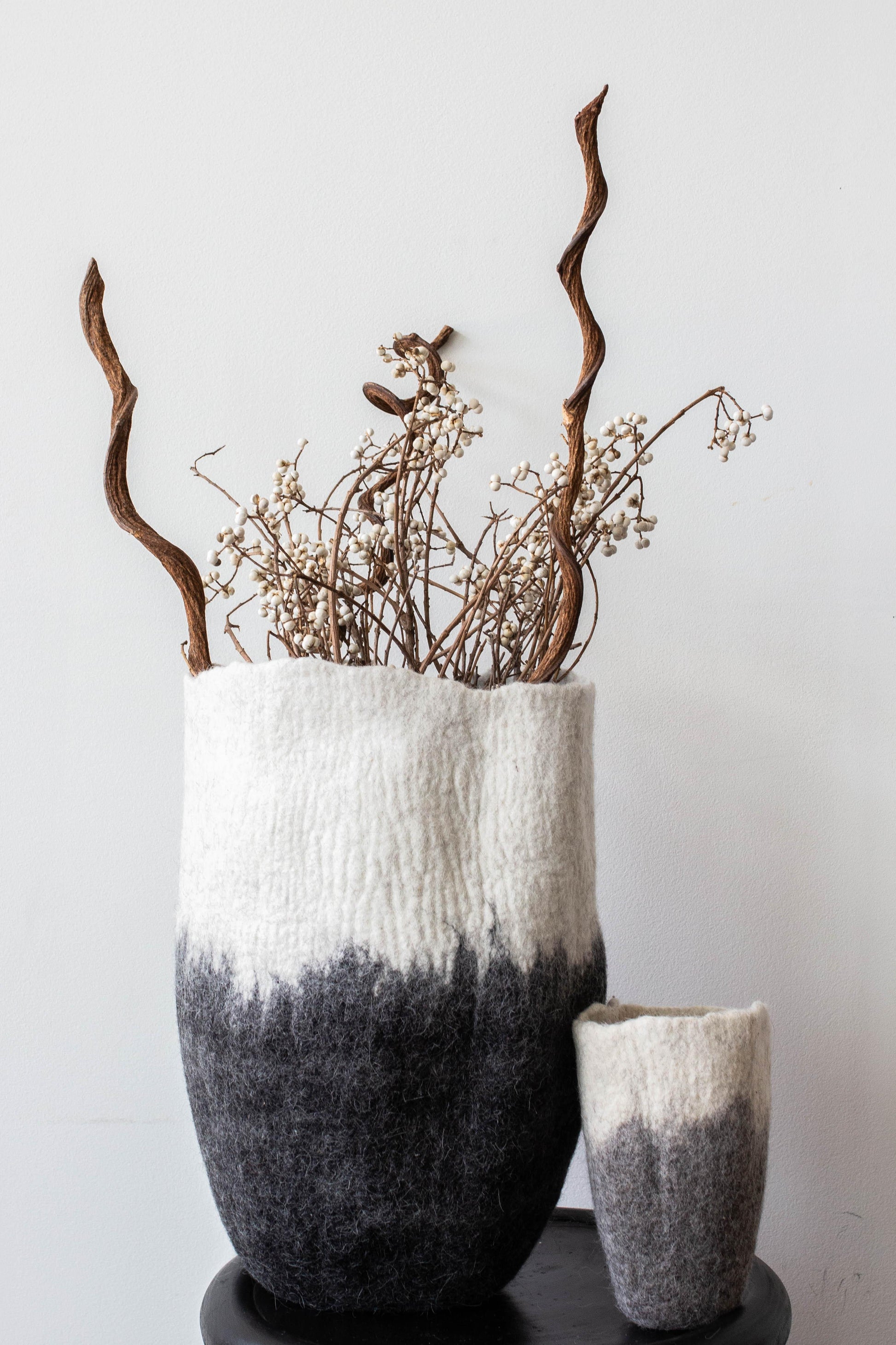 Large white to charcoal ombré felted wool floor vase styled with dried branches and wildflowers on black pedestal against white wall

