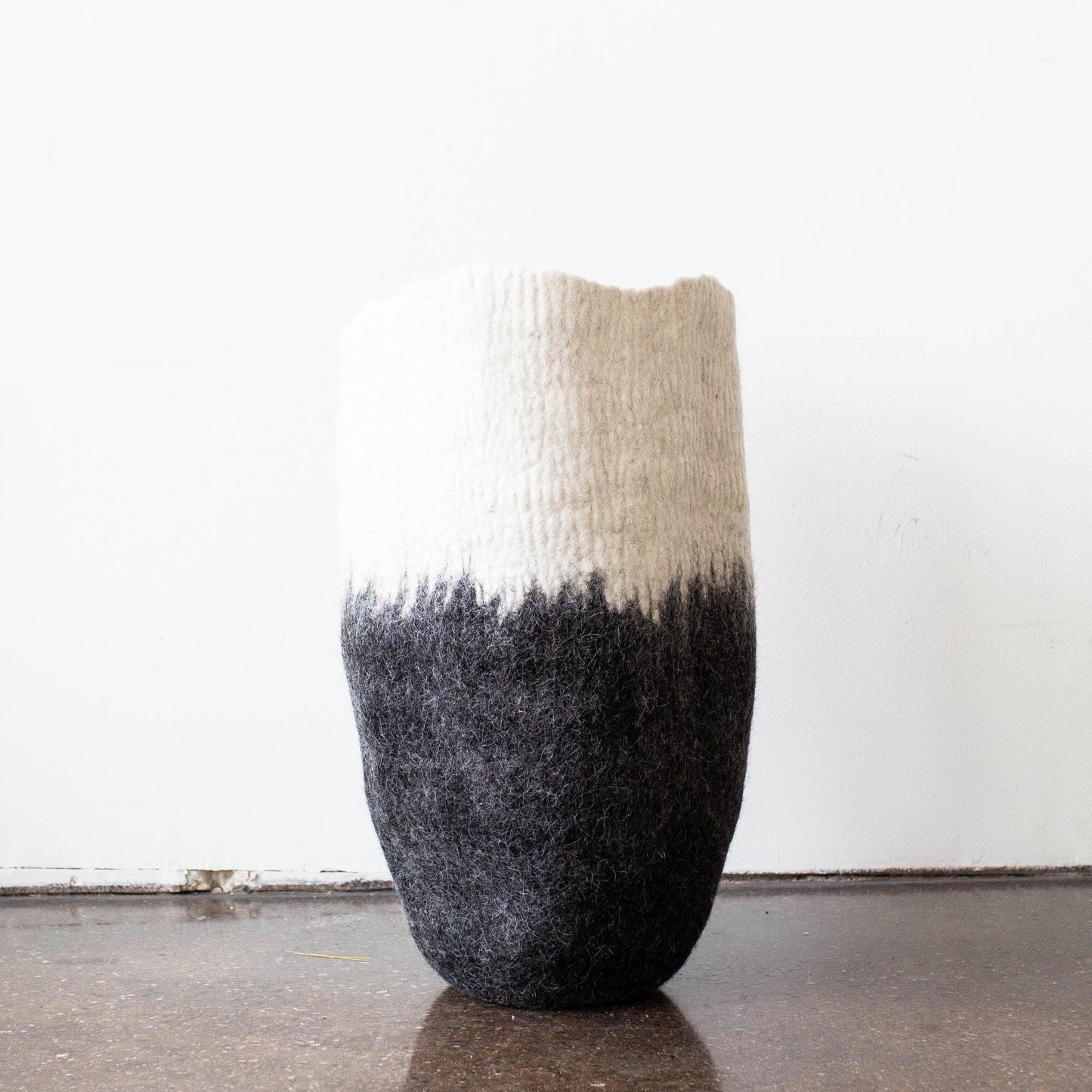 Tall white to charcoal ombré felted wool floor vase standing solo on dark wood floor against white wall showing full sculptural silhouette

