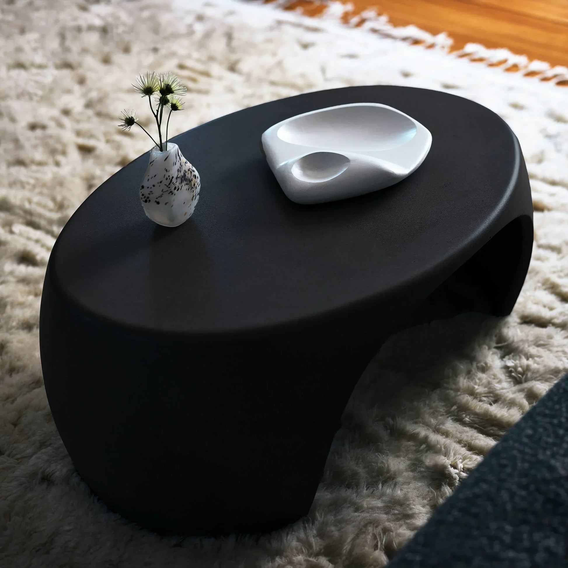Black Nook concrete coffee table styled with speckled ceramic vase and sculptural white dish on ivory shag rug

