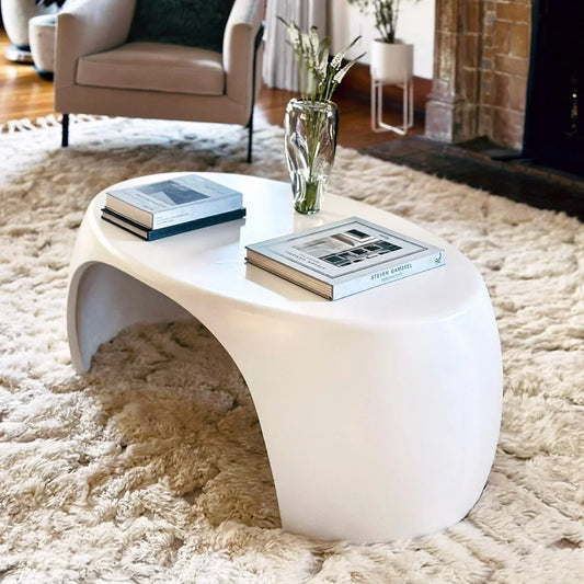 White Nook concrete coffee table styled with books and glass vase on ivory shag rug beside leather chair and fireplace

