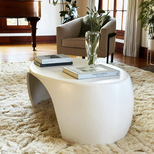 White Nook concrete coffee table styled with art books and glass vase on ivory shag rug beside taupe chair and grand piano

