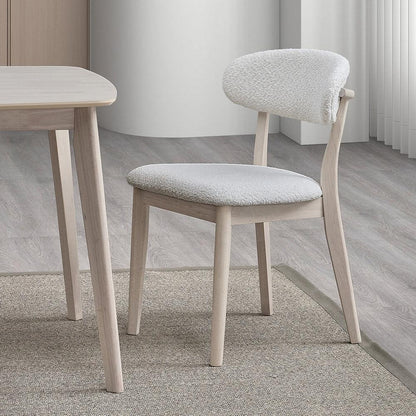 White-washed Scandinavian dining chair with bouclé upholstered seat and curved backrest shown beside matching table leg on neutral rug

