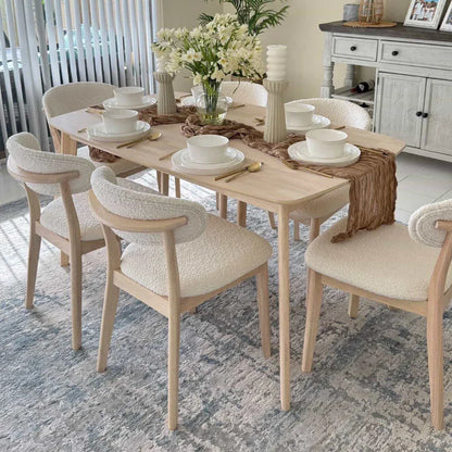 White-washed Scandinavian wood dining set styled with white place settings woven table runner fresh florals and bouclé chairs on a blue vintage rug

