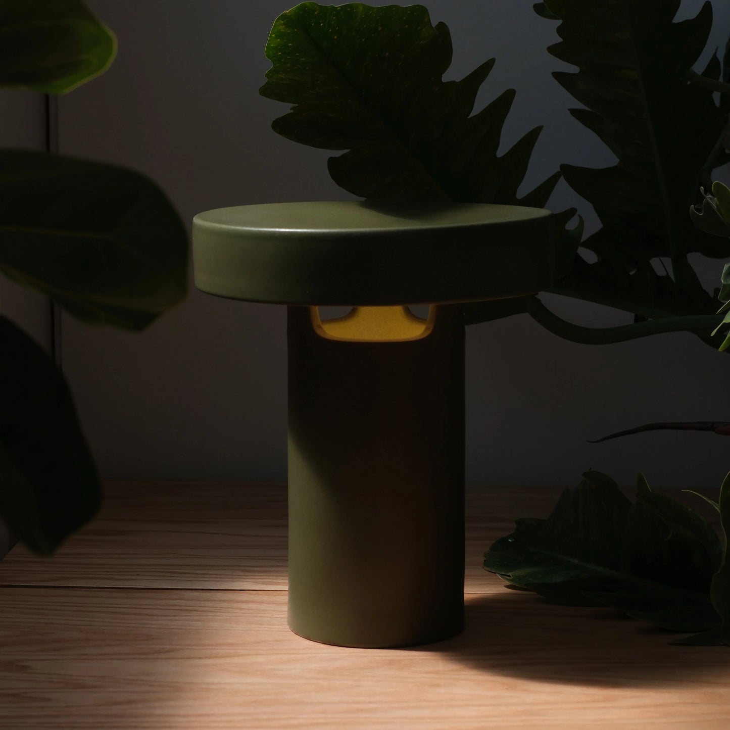 Moss green ceramic slot table lamp glowing warm amber light on wood surface surrounded by dark tropical foliage

