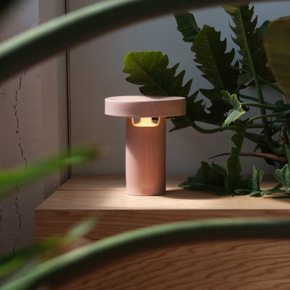 Pink glossy ceramic slot table lamp glowing warm light on wood shelf surrounded by tropical green plants

