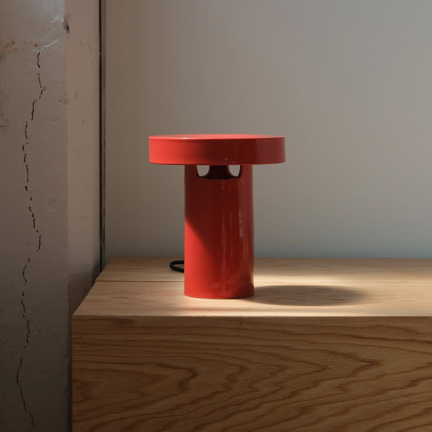Red glossy ceramic slot table lamp casting warm downward glow on wood surface against neutral wall in minimalist interior

