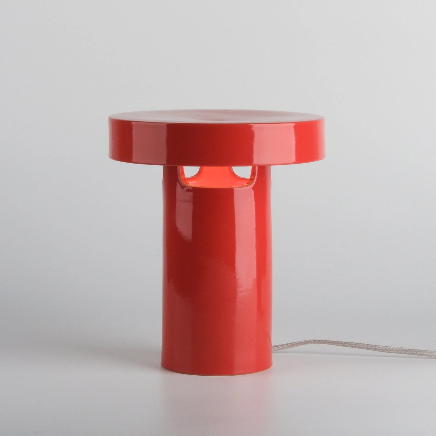 Red glossy ceramic slot table lamp with three slot openings on white background showing full cylindrical form and fabric cord

