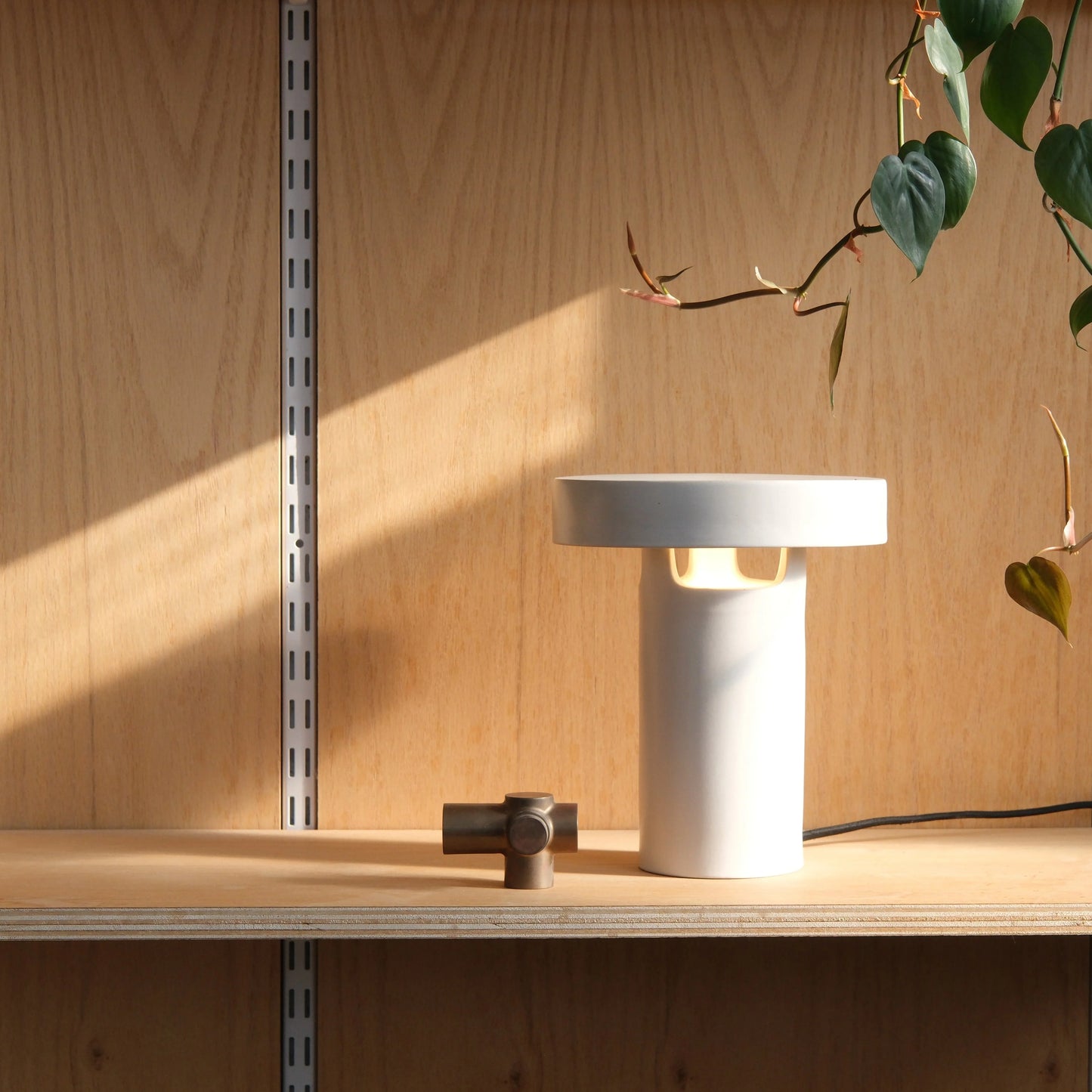 White glossy ceramic slot table lamp glowing warm light on wood shelf with fabric cord dimmer and trailing green plant


