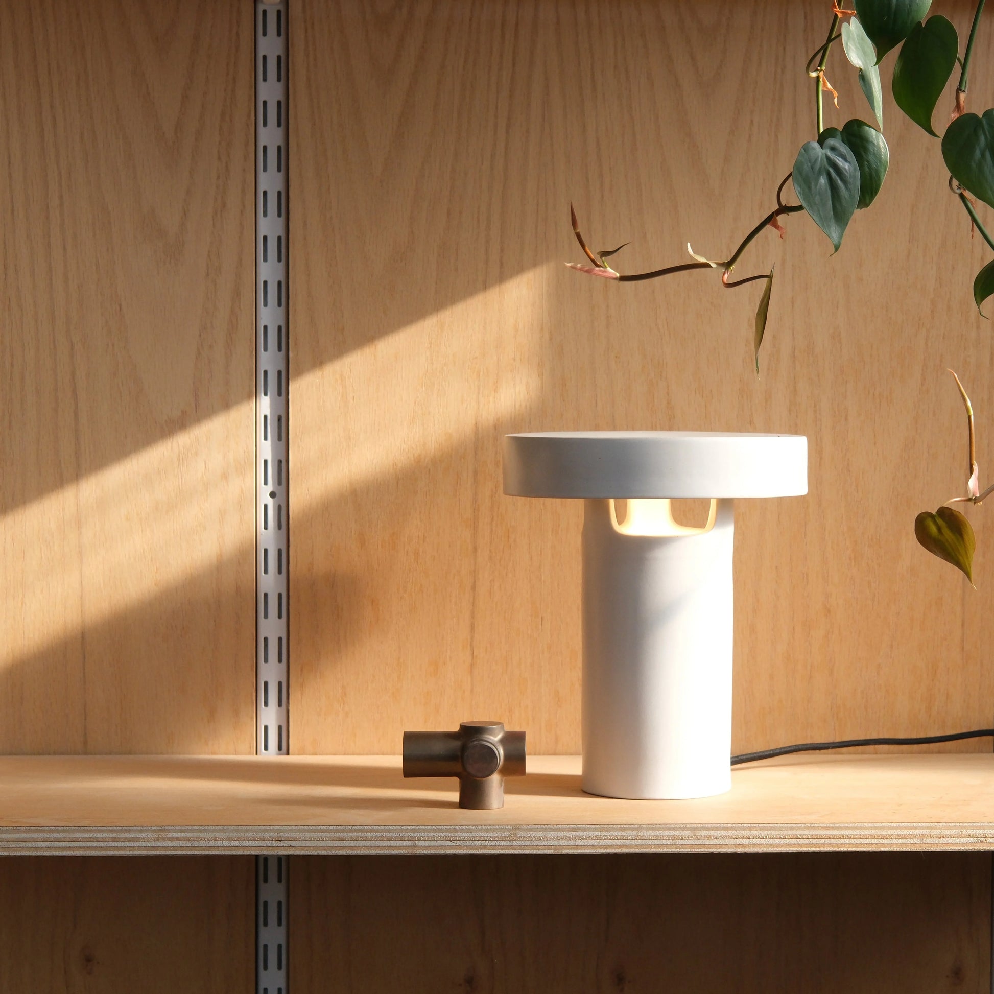 White glossy ceramic slot table lamp glowing warm light on wood shelf with fabric cord dimmer and trailing green plant

