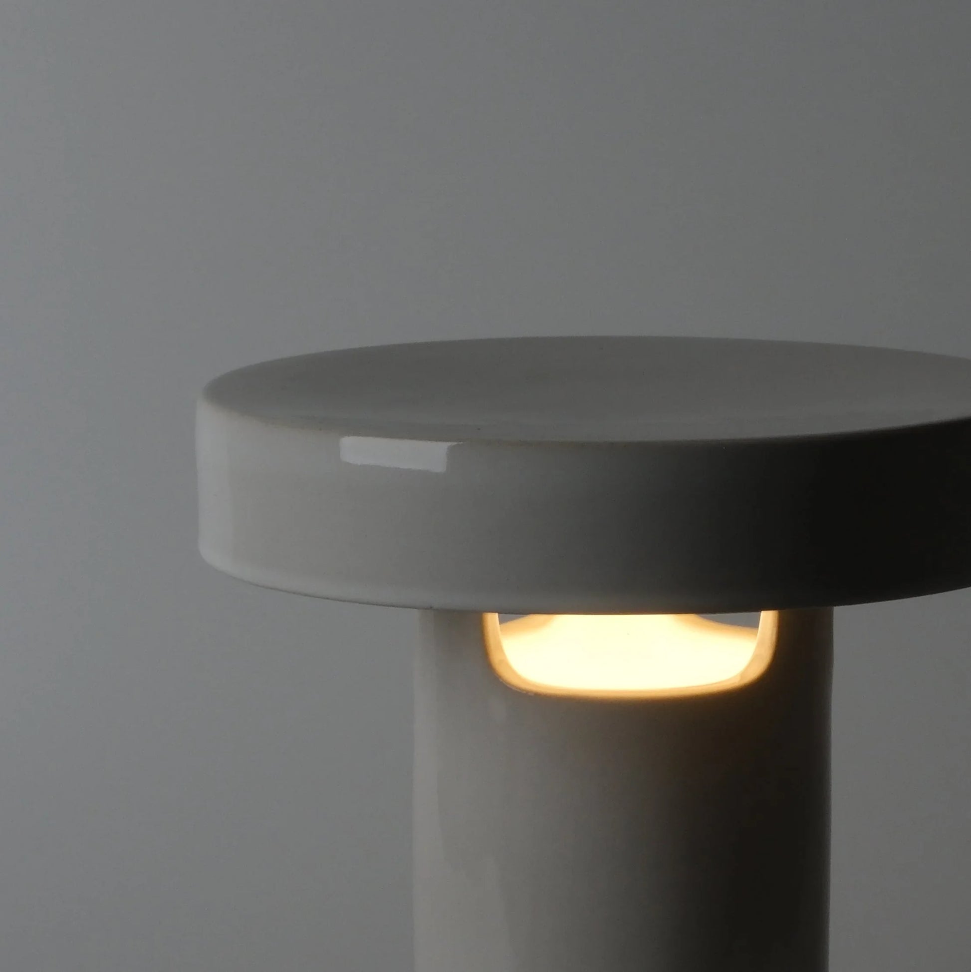 Close-up of white glossy ceramic slot table lamp showing warm LED glow through single slot opening on gray background

