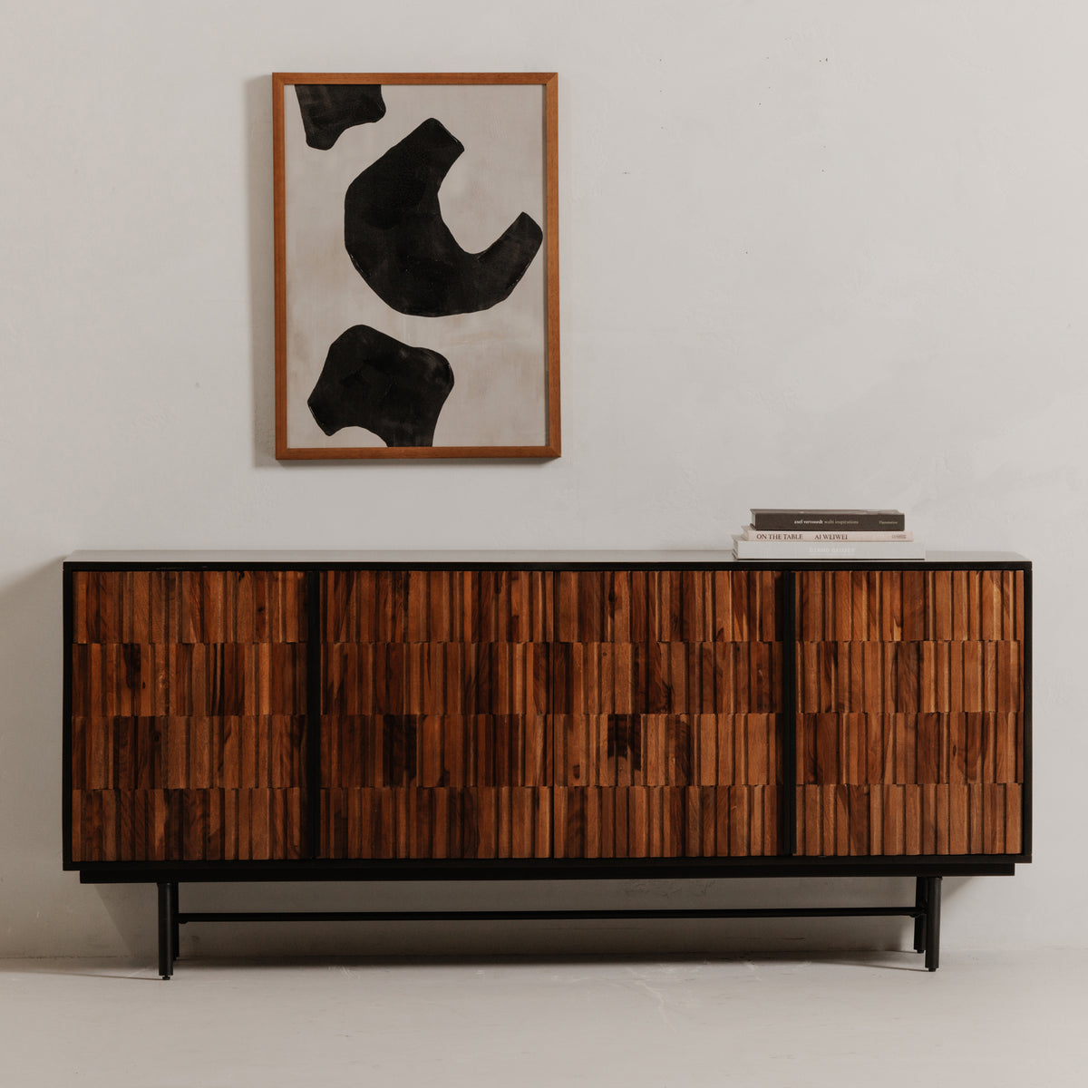 Solid sheesham and mango wood sideboard with textured brown doors and a black iron base, styled beneath abstract wall art with books on top.