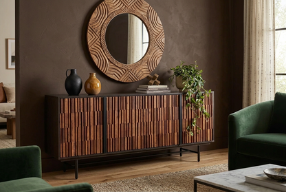 Solid sheesham and mango wood sideboard with textured brown doors and a black iron base, styled with vases, books, and a trailing plant beneath a round mirror.