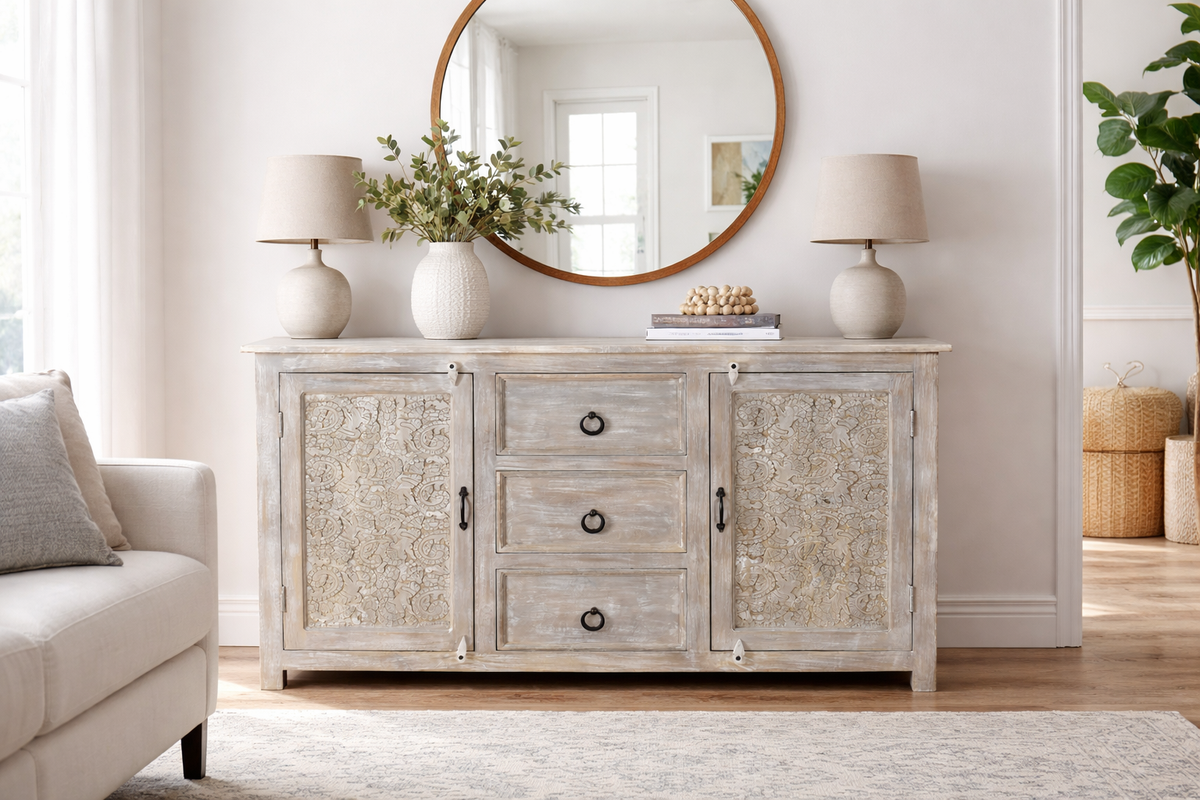 Hand carved antique sideboard with both doors closed showing full floral carved panel detail three center drawers with iron ring pulls white antique finish styled with round mirror neutral vases and linen lamps