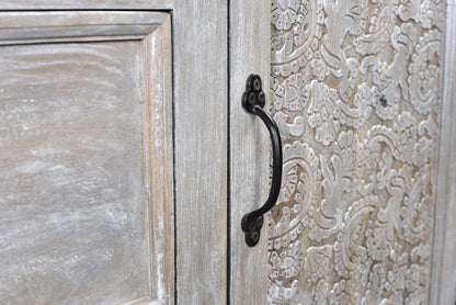 Close up of hand carved antique sideboard showing black iron pull handle against detailed floral carved door panel and smooth reclaimed acacia wood door frame in white antique finish