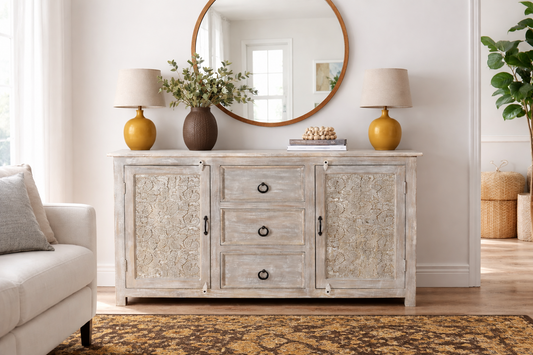 Hand carved antique sideboard in white antique finish styled in living room with round wood mirror two yellow ceramic lamps dark vase with greenery and floral area rug
