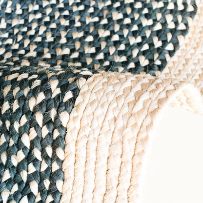 Angled edge detail of round sisal rug showing curved border transition between charcoal black two-tone braided field and natural cream border braid with individual sisal fiber strands visible