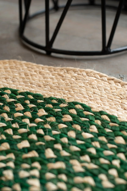 Low angle lifestyle detail of round sisal rug forest green variant on concrete floor showing curved edge braid transition from natural cream border to deep forest green two-tone field with black metal stool base in background