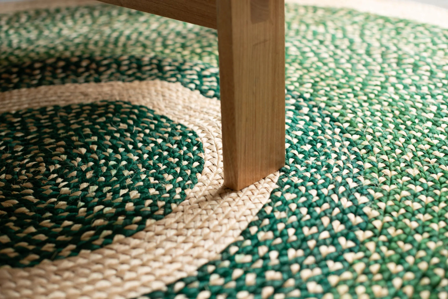 Overhead lifestyle detail of round sisal rug forest green variant showing concentric cream ring motif and deep forest green two-tone braided field with natural ash wood furniture leg resting on cream border ring