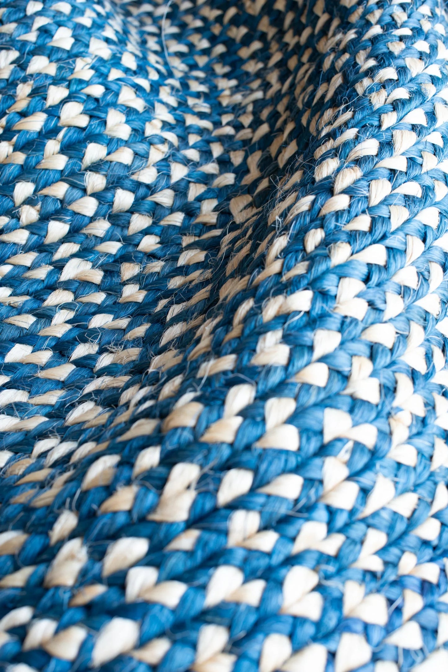 Angled close-up of round sisal rug marine blue variant showing curved concentric braid rings with deep marine blue and natural cream two-tone weave pattern and individual sisal fiber texture throughout