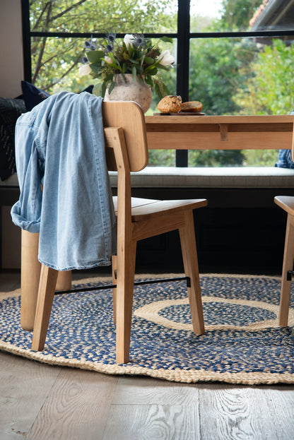 Large round sisal rug marine blue variant styled under natural ash dining table and chair with denim jacket draped over chair back flower vase and garden view through black steel windows on wood plank floor