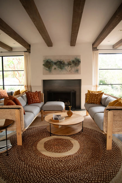Large round sisal rug red earth variant anchoring living room seating area with exposed wood beam ceiling fireplace and warm terracotta pillows