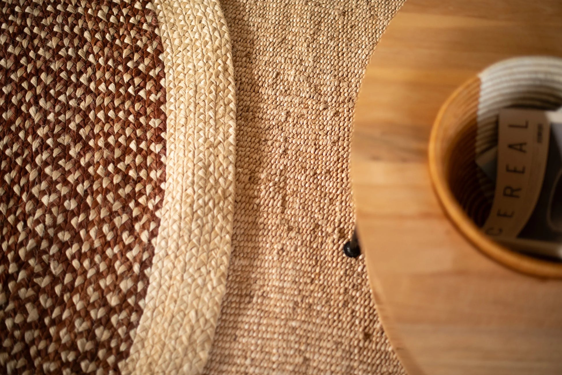 Overhead lifestyle detail of round sisal rug red earth variant showing curved cream ring motif separating dusty mauve braided field from natural sisal border with round wood tray and rolled magazine in foreground