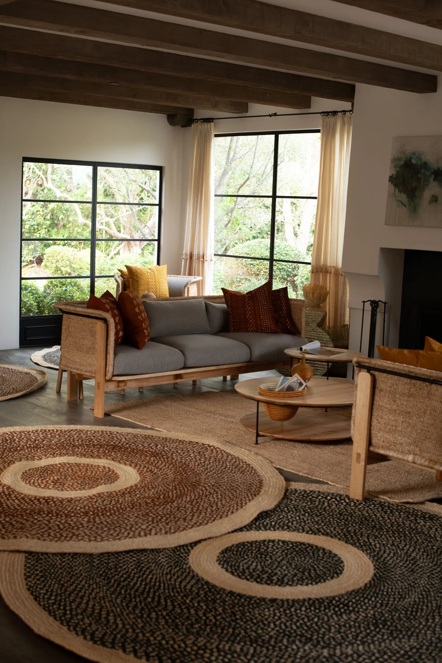 Large round sisal rugs black and red earth variants lifestyle living room with two rattan chairs grey sofa terracotta rust pillows exposed wood beam ceiling black steel windows and garden view