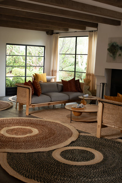 Large round sisal rugs black and red earth variants lifestyle living room with two rattan chairs grey sofa terracotta rust pillows exposed wood beam ceiling black steel windows and garden view