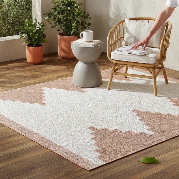 Hauteloom Djugun Peach Fuzz Aztec geometric rug on a wood patio with a rattan chair, side table, and plants.