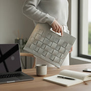 Woman holds Glacial Gray Artisan Woven Leather Laptop Clutch in home office with laptop, coffee, and notebook on wooden desk.