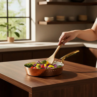 Copper frying pan with brass handle, filled with colorful vegetables, being stirred by a woman's hand in a sunlit kitchen.