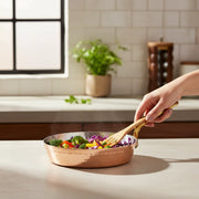 Copper frying pan with brass handle on concrete countertop, person stirring colorful vegetables with a wooden spatula.