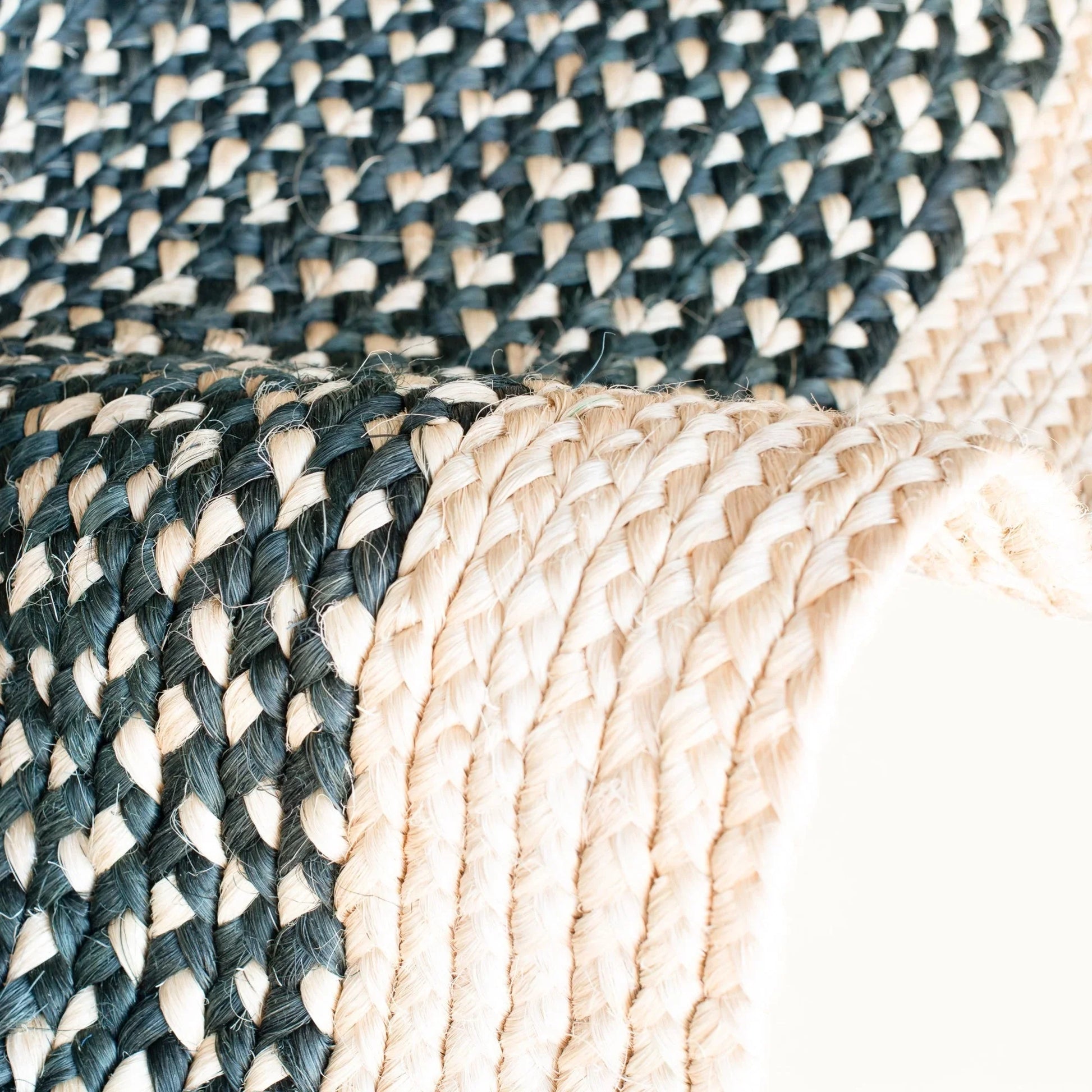 Corner detail of medium round sisal rug black variant showing raw fringe edge finishing and deep forest green two-tone braided weave on white background