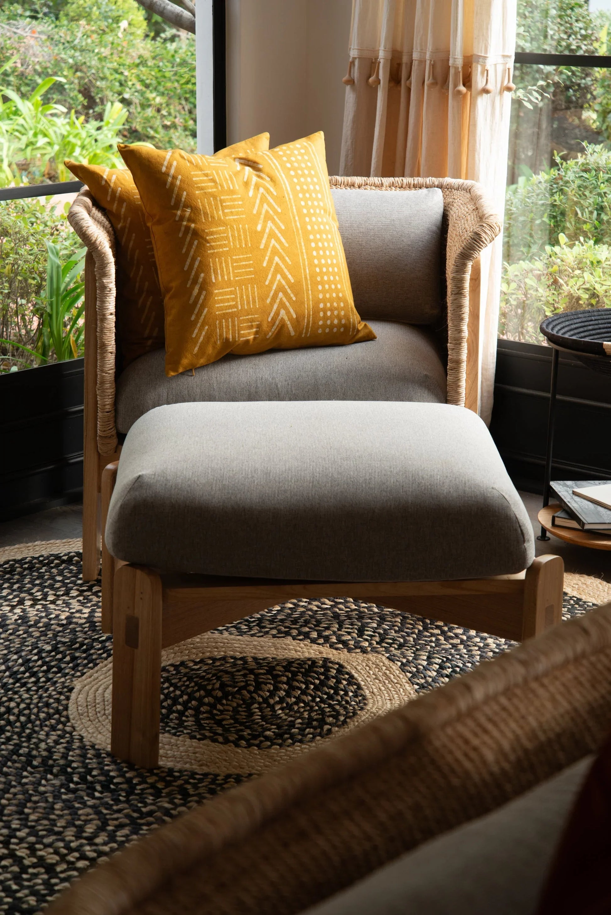 Handwoven medium round sisal rug black variant styled under rattan armchair with ottoman grey cushion mustard mud cloth pillows and lush garden view through black steel windows