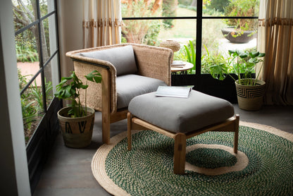 Handwoven medium round sisal rug forest green variant anchoring rattan armchair and ottoman with open book woven plant baskets and lush garden view through black steel windows on concrete floor