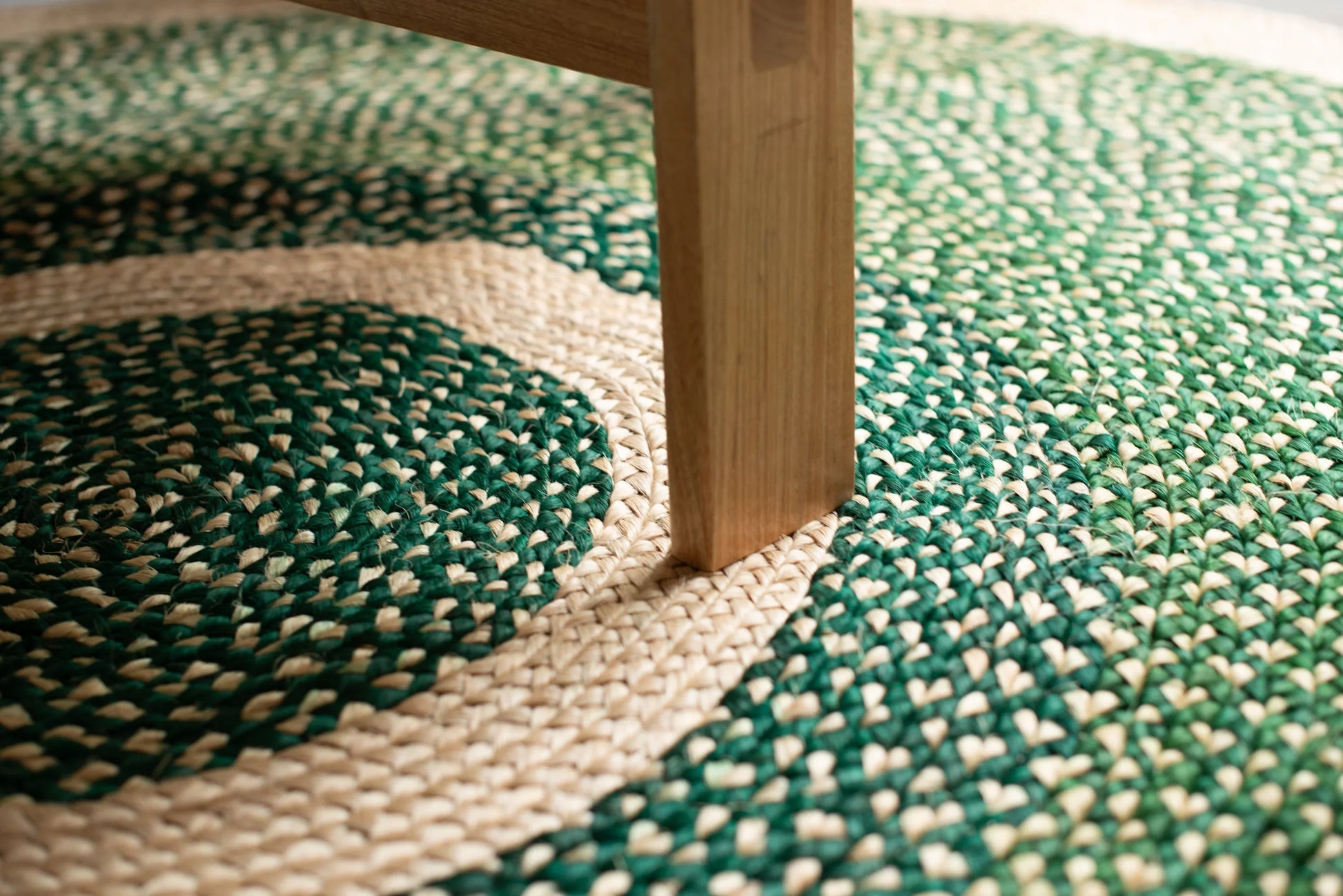 Overhead detail of medium round sisal rug forest green variant showing concentric cream ring motif and deep forest green two-tone braided field with natural ash wood furniture leg resting on cream border ring