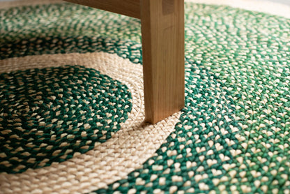 Overhead detail of medium round sisal rug forest green variant showing concentric cream ring motif and deep forest green two-tone braided field with natural ash wood furniture leg resting on cream border ring