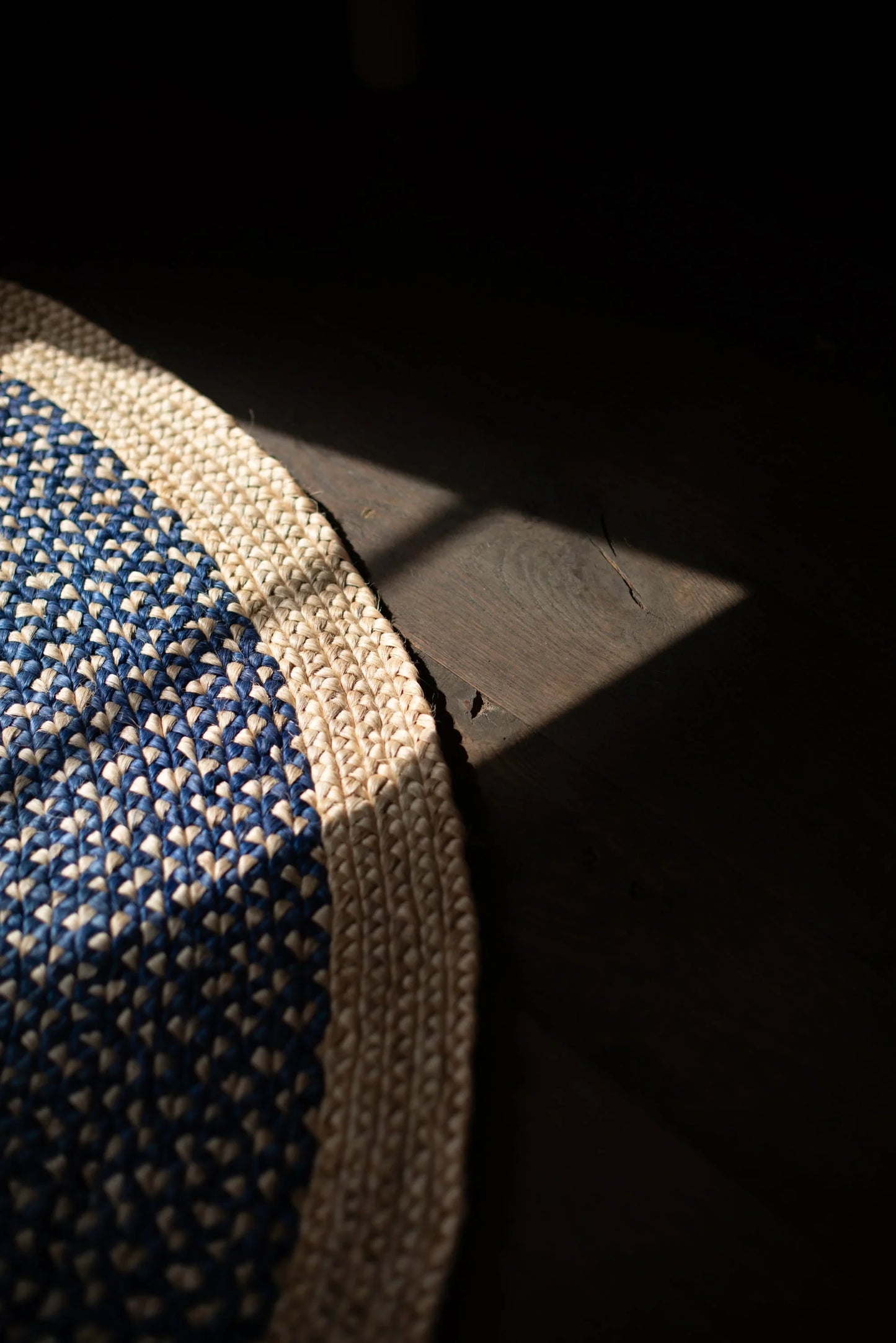 Curved edge detail of medium round sisal rug marine blue variant showing deep marine blue two-tone braided field meeting natural cream border braid with dramatic raking shadow on dark concrete floor