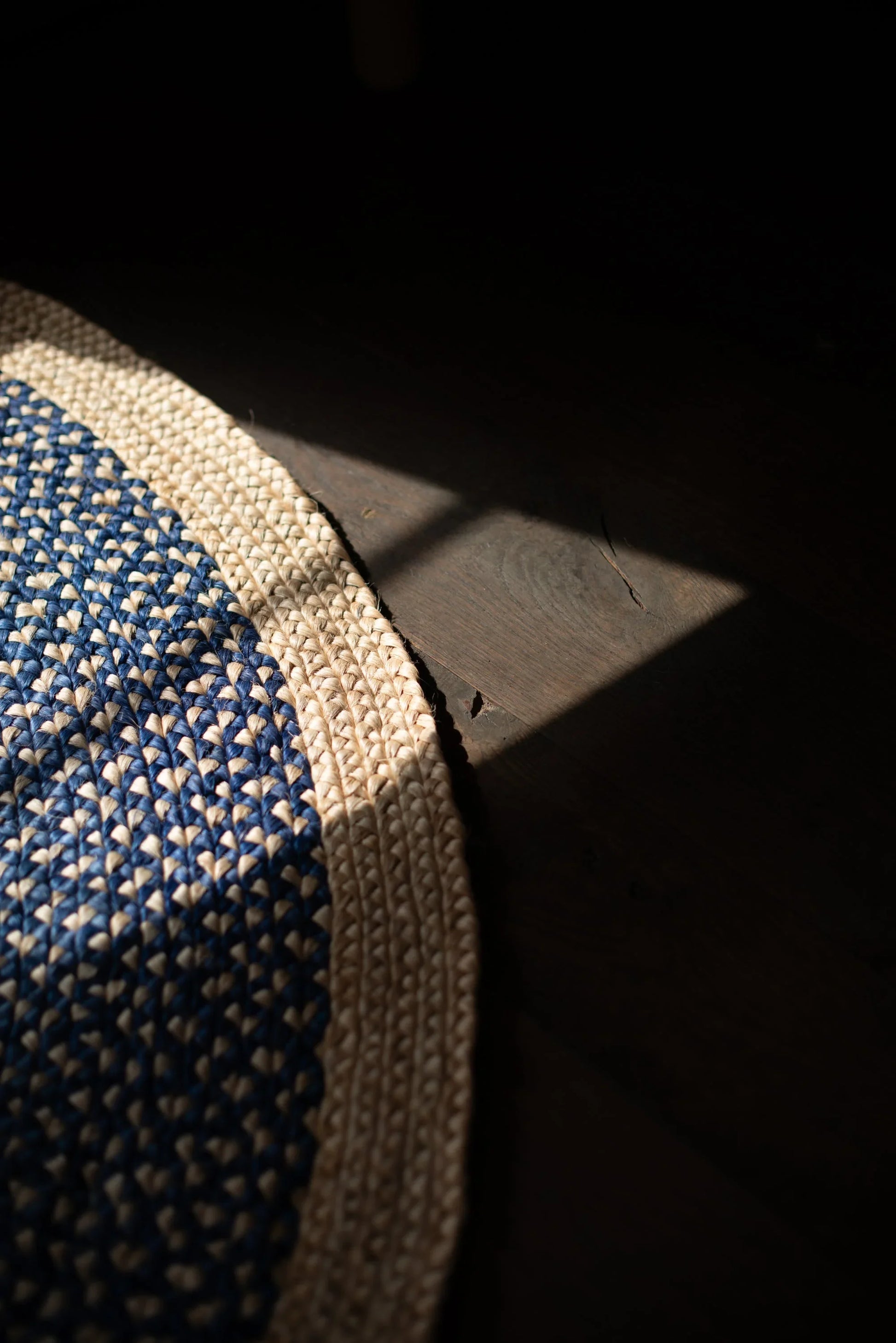 Curved edge detail of medium round sisal rug marine blue variant showing deep marine blue two-tone braided field meeting natural cream border braid with dramatic raking shadow on dark concrete floor