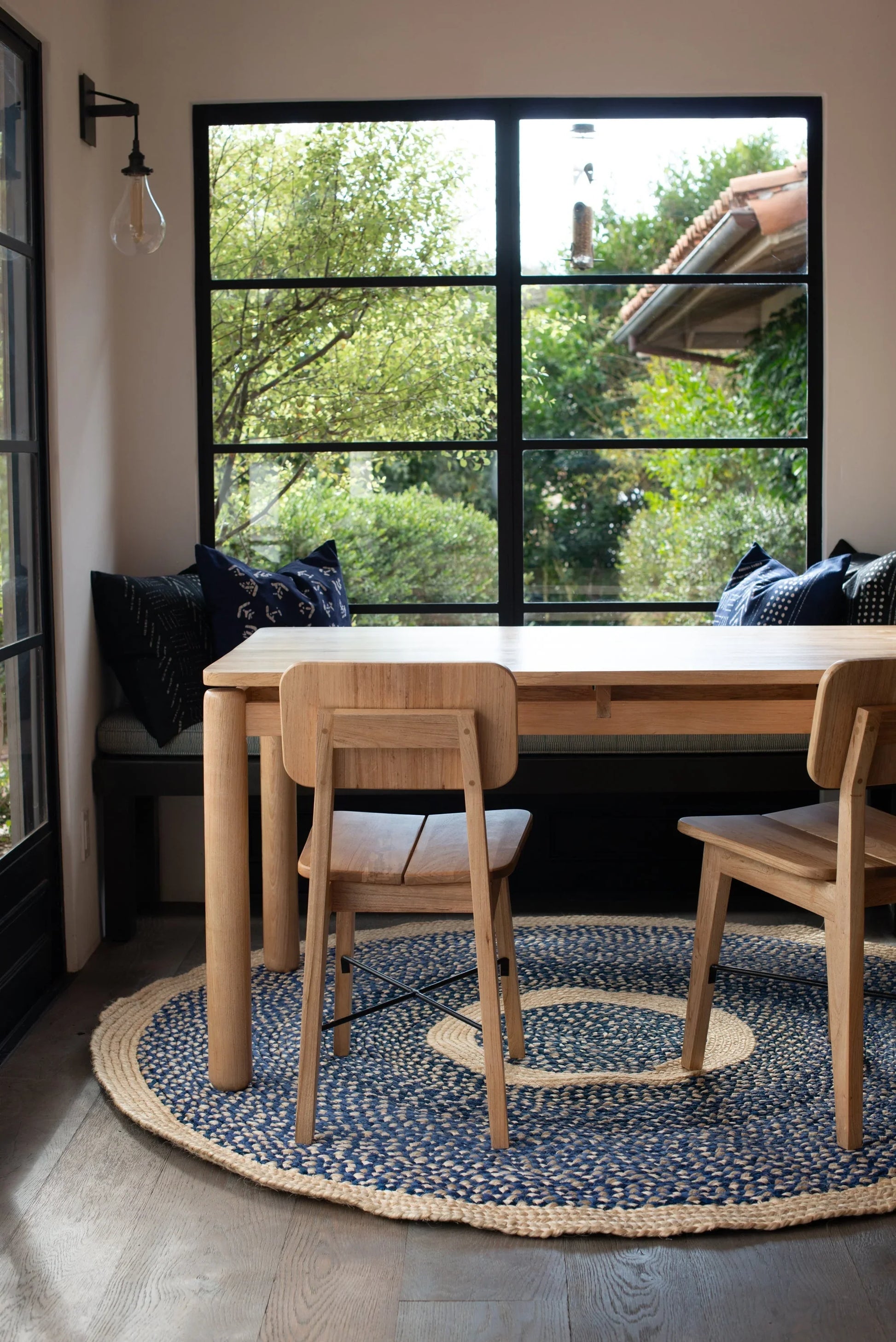 Handwoven medium round sisal rug marine blue variant anchoring natural ash dining table and chairs with navy bench cushion indigo pillows and lush garden view through black steel windows on wood plank floor