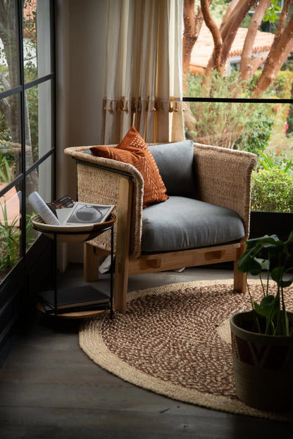 Handwoven medium round sisal rug red earth variant styled under rattan armchair with grey cushion terracotta pillow black side table and lush garden view through black steel windows