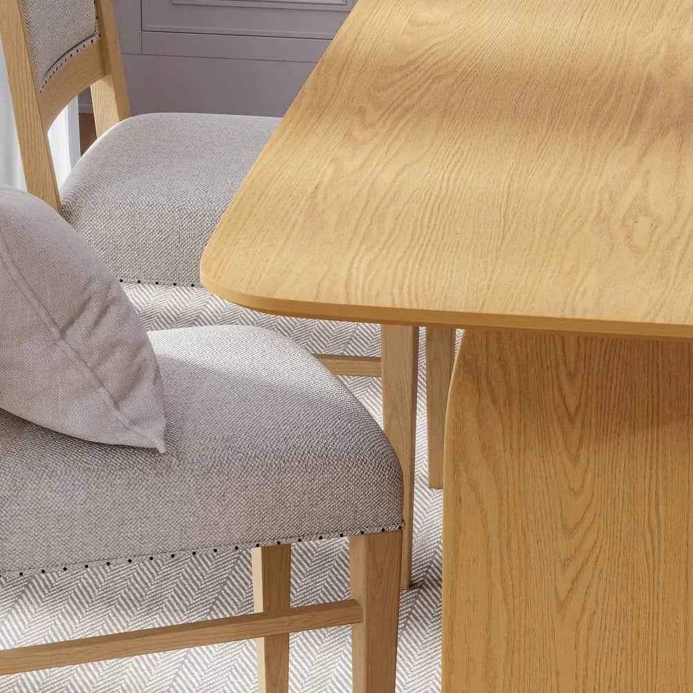 Close up view highlighting a gray upholstered dining chair with nailhead trim positioned next to a natural solid oak double pedestal dining table tabletop edge