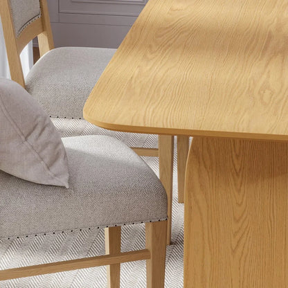 Close up view highlighting a gray upholstered dining chair with nailhead trim positioned next to a natural solid oak double pedestal dining table tabletop edge