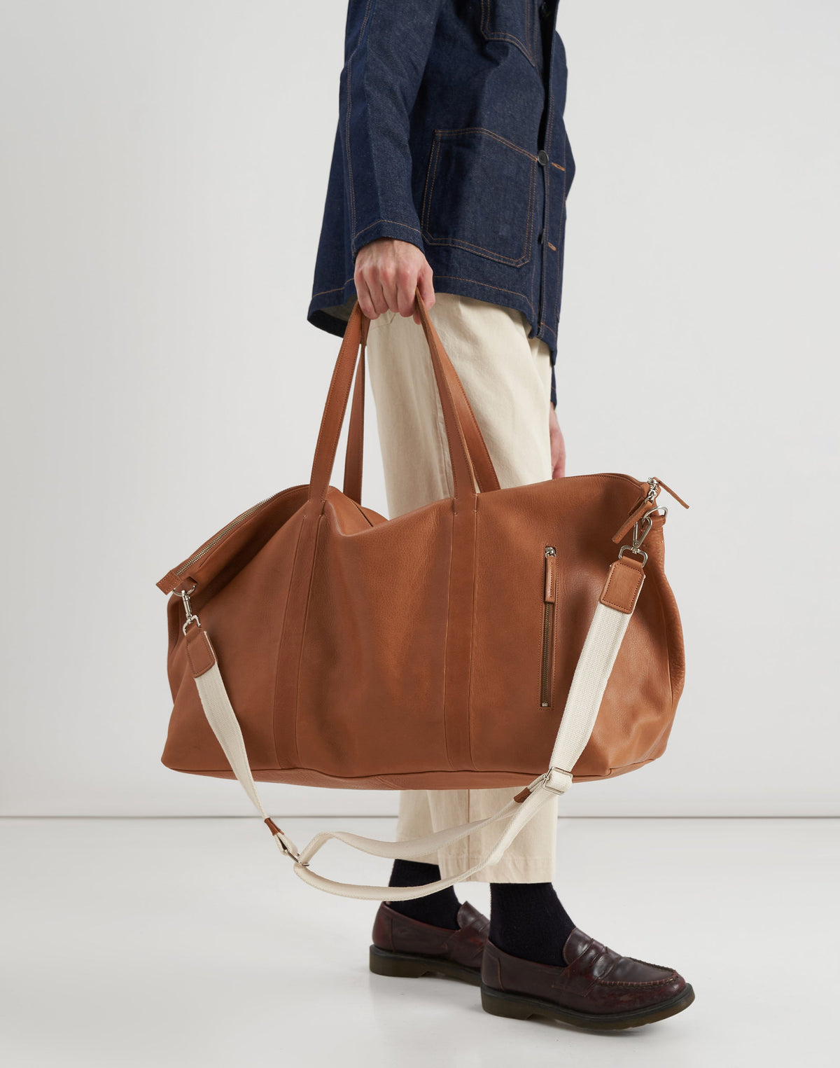 Model carrying the Malbec leather weekender in Tobacco by its twin handles, with the cream shoulder strap hanging below.
