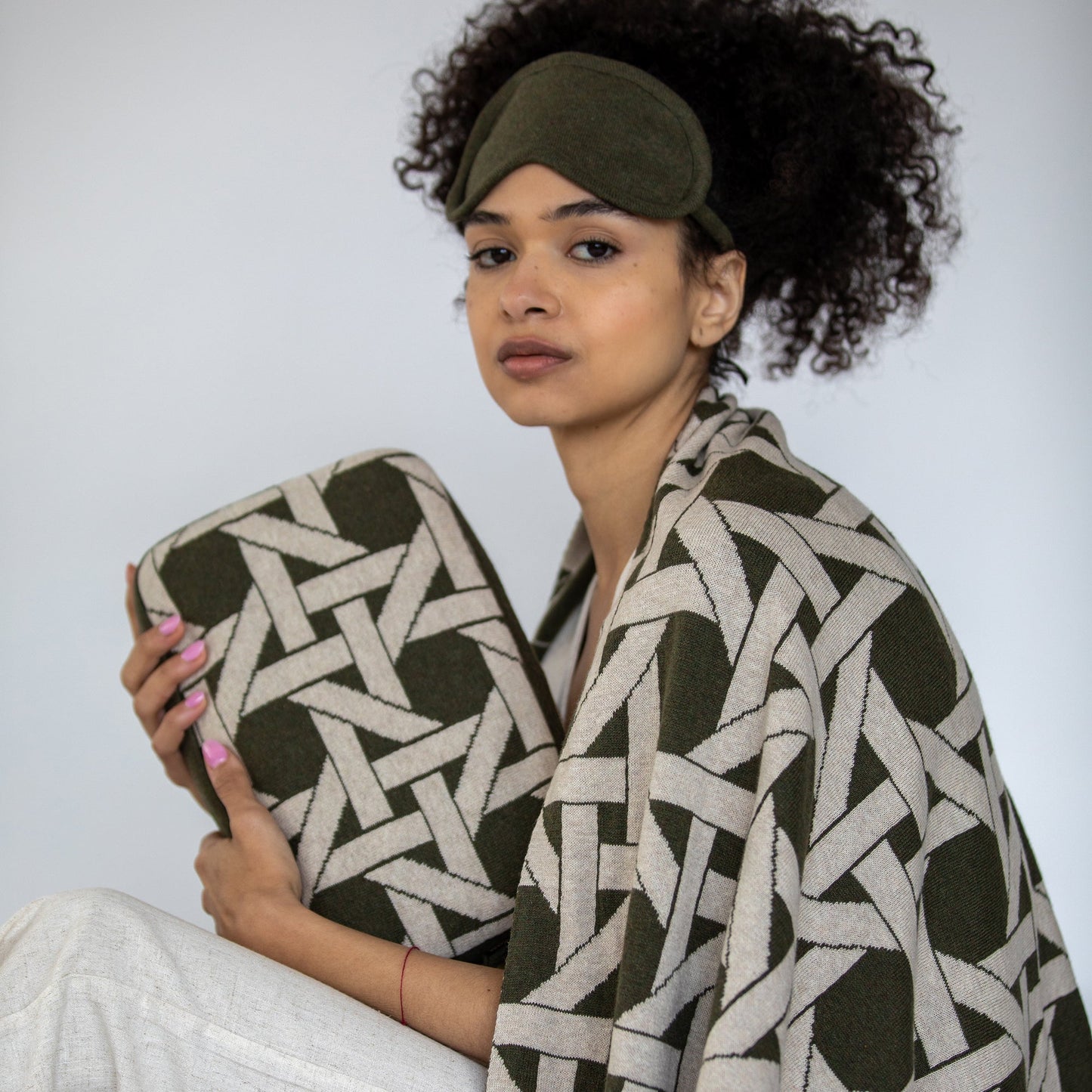 Woman wearing olive green organic cotton travel blanket draped over shoulders with matching eye mask and geometric compact pouch