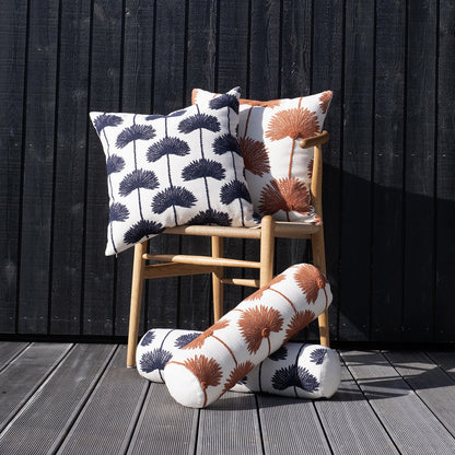 Palm leaf embroidered throw pillows and bolster cushions both navy off white and sienna off white variants propped against natural rattan chair on grey wood deck with black timber wall in bright sunlight